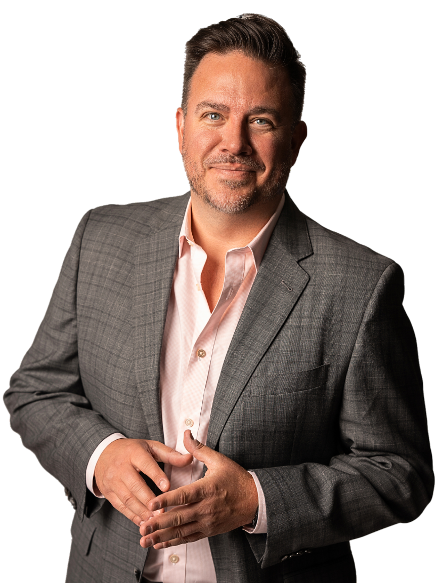 A confident man wearing a gray suit jacket over a light pink dress shirt, standing against a plain white background, with a slight smile and folded hands. Nick Sundahl, Realtor - Lakes | Sotheby's International Realty
