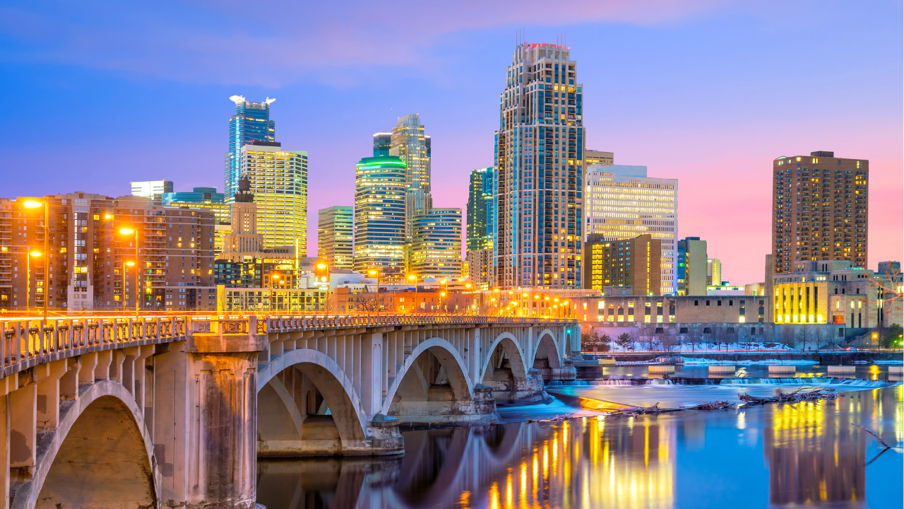 City skyline with illuminated skyscrapers and a bridge over a river, vibrant sunset in the background, reflections on the water. Twin Cities Relocation with Nick Sundahl