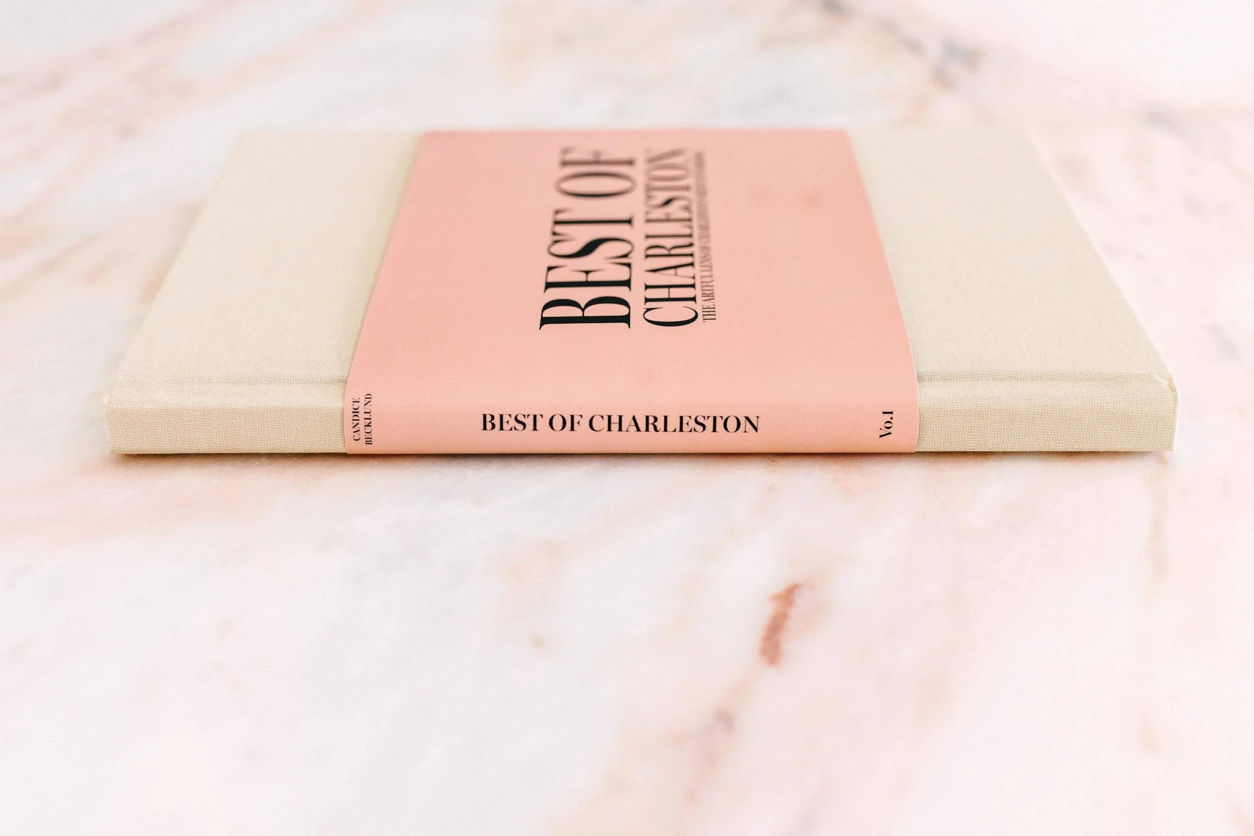 A beige book with a pink cover titled 'Best of Charleston'. The book is lying flat on a marble surface, displaying the title and volume number.