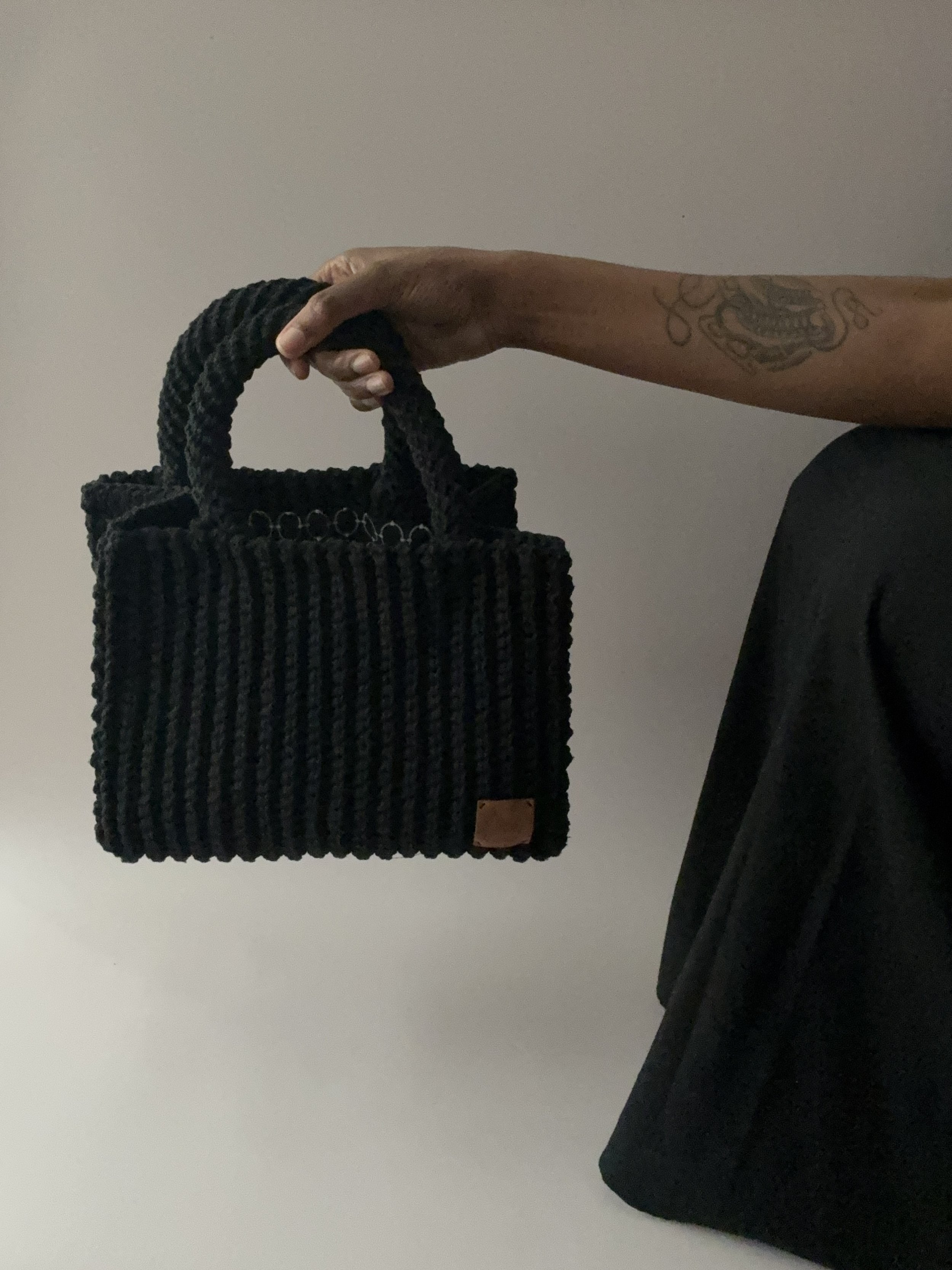 Main tenant une sac noir en crochet, tenue d'une personne portant une chemise noire, avec un tatouage sur le bras, contre un mur clair.