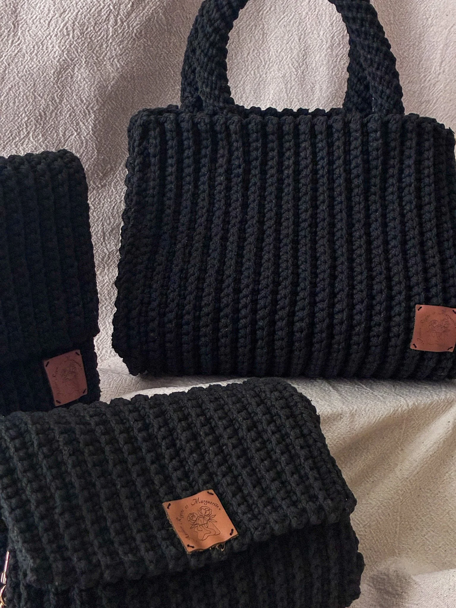 Trois sacs à main noirs en crochet avec des étiquettes en cuir, posés sur une surface de tissu beige.