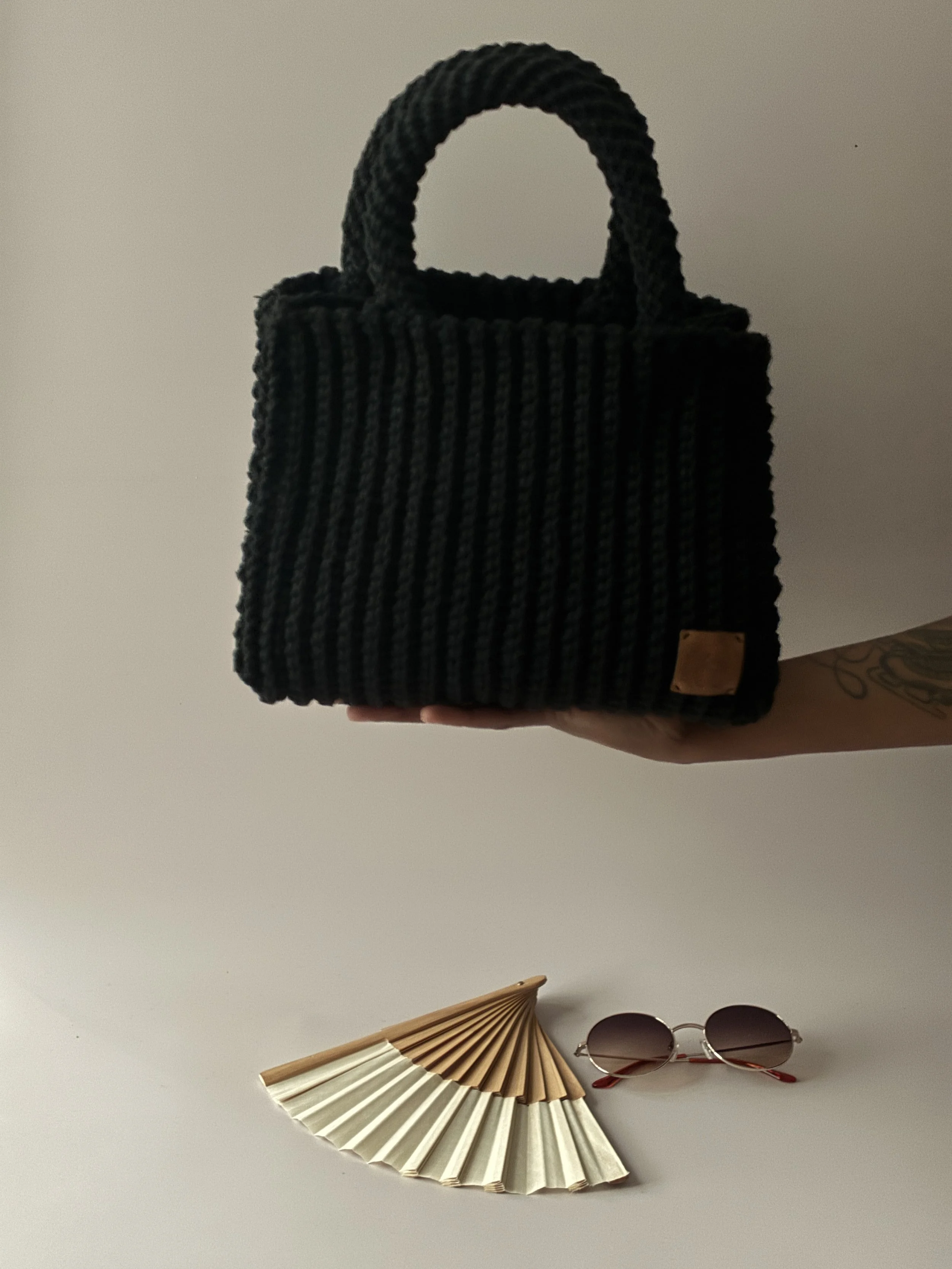 Une main tient un sac noir en tricot avec une poignée à la main, à côté se trouvent des lunettes de soleil rondes et un éventail en bois et papier beige et blanc.