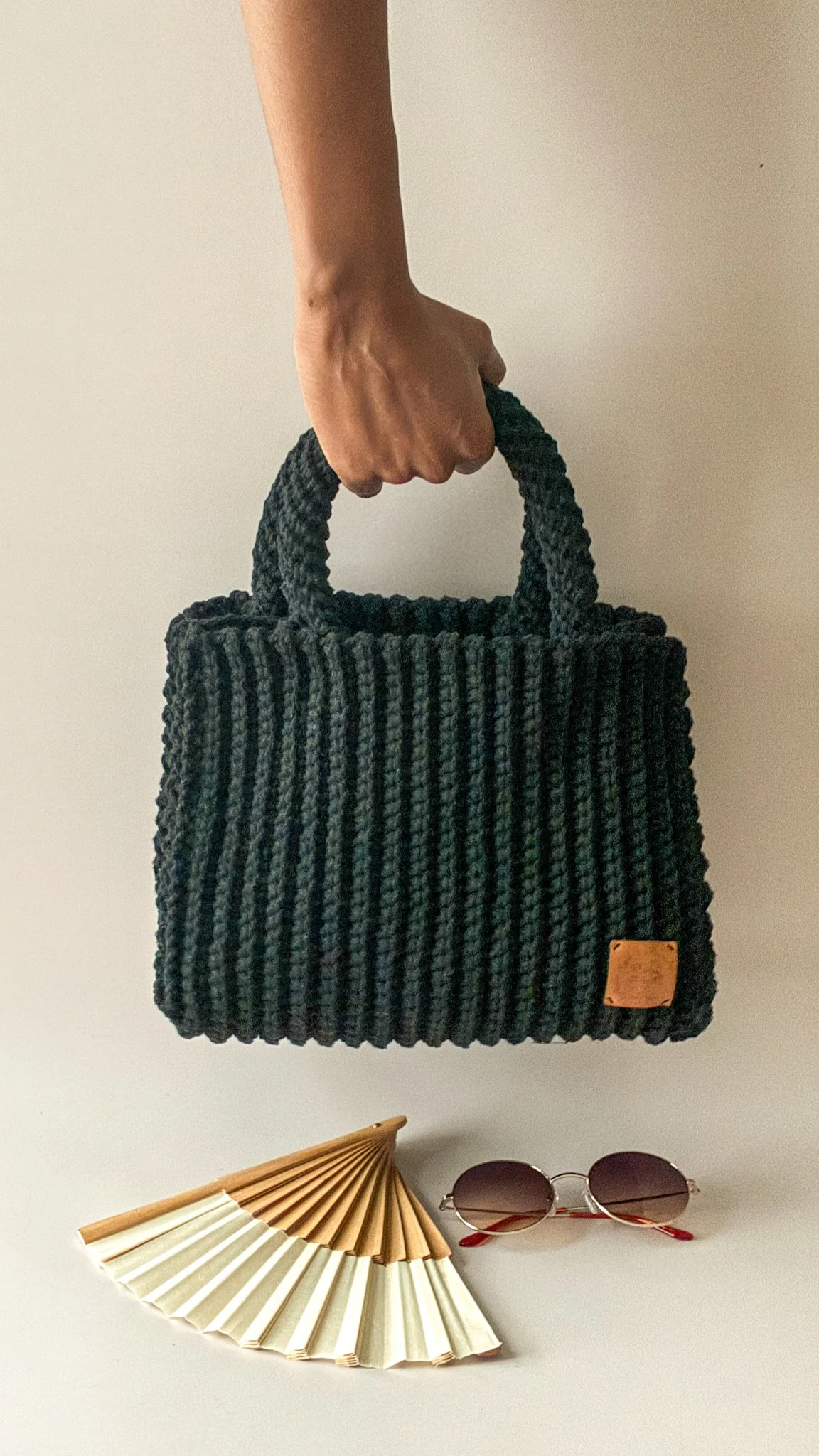 Une main tenant un sac à main noir en tricot avec une étiquette en cuir en bas à droite, une paire de lunettes rondes et un éventail blanc et beige sur une surface blanche.