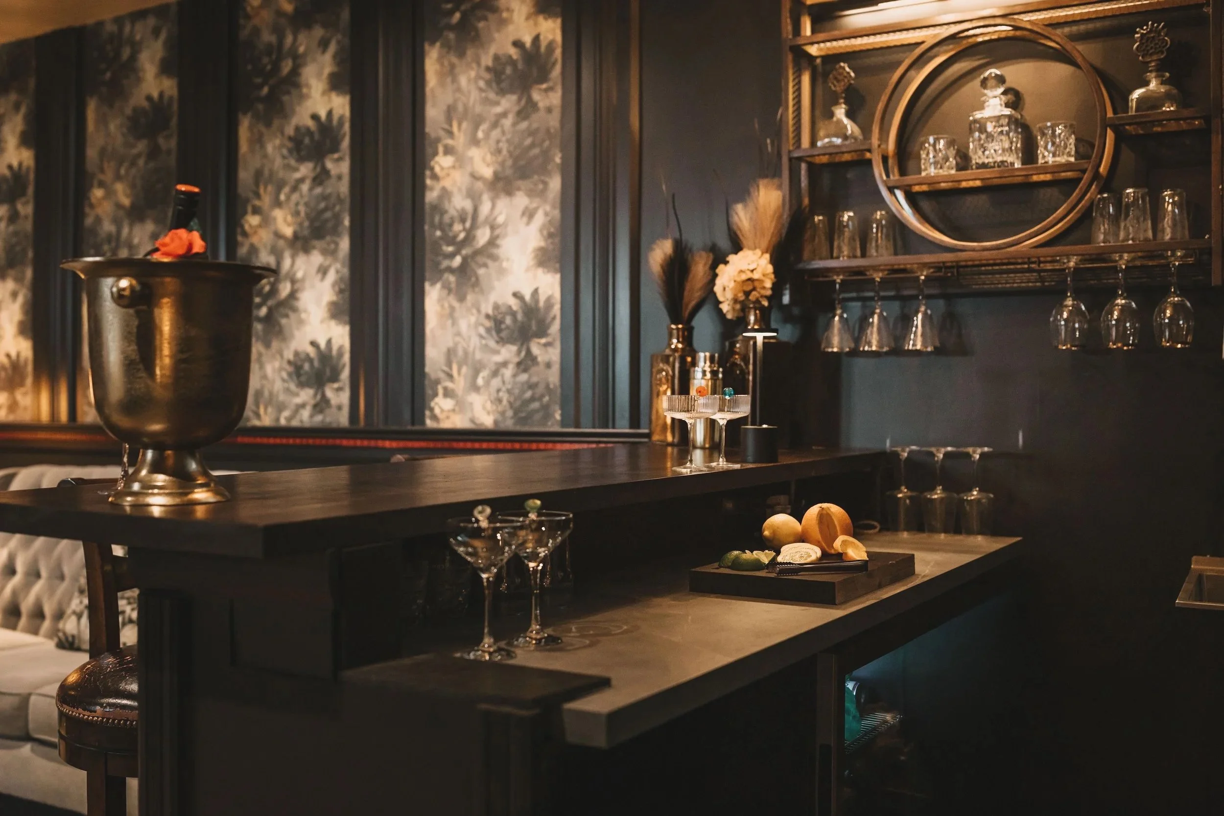 A dark wooden bar counter with a metallic ice bucket, cocktail glasses, and a small tray of cheeses and limes. Behind the bar is a wall with dark floral wallpaper and decorative glassware on shelves.