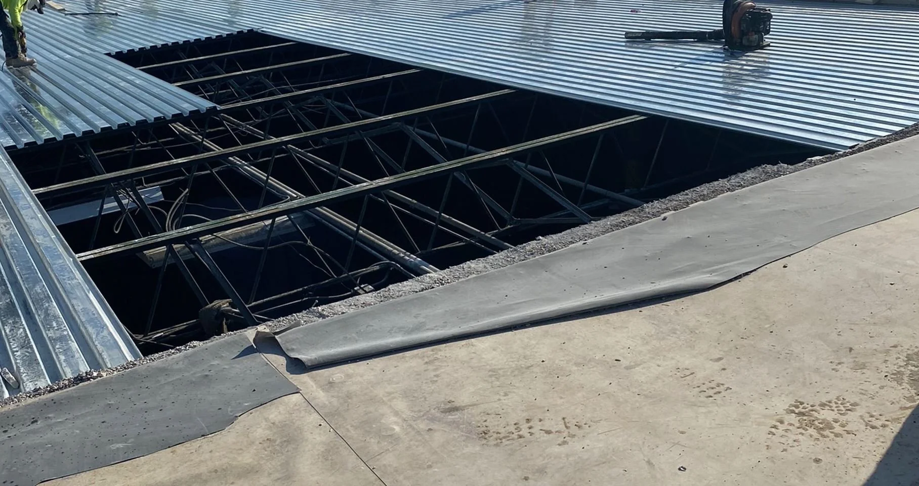 Construction site showing metal framework and roofing panels being installed over an open space. Concrete surface in the foreground.