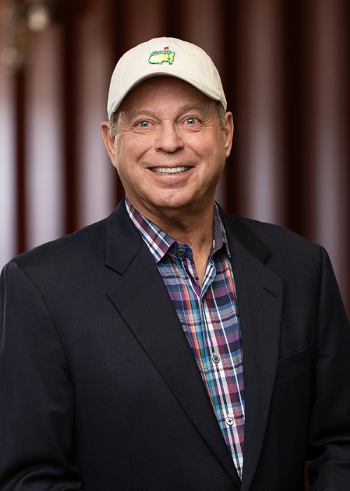 Larry Johnson wearing a beige hat with a crocodile logo, a dark blazer, and a colorful plaid shirt, standing in front of a brown curtain.