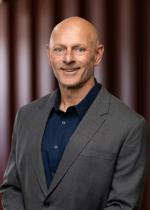 Nick Gignac, wearing a dark blue shirt and a gray blazer, smiling in front of a blurred brown curtain background.