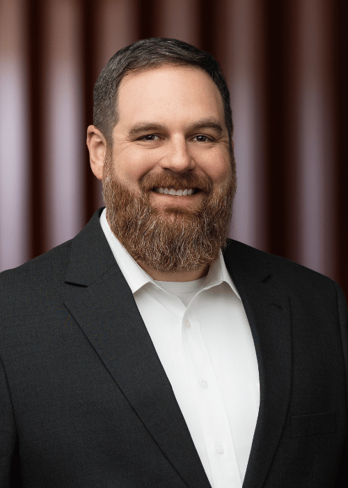 John Jamison, wearing a suit and white shirt, smiling in front of a brown curtain background.