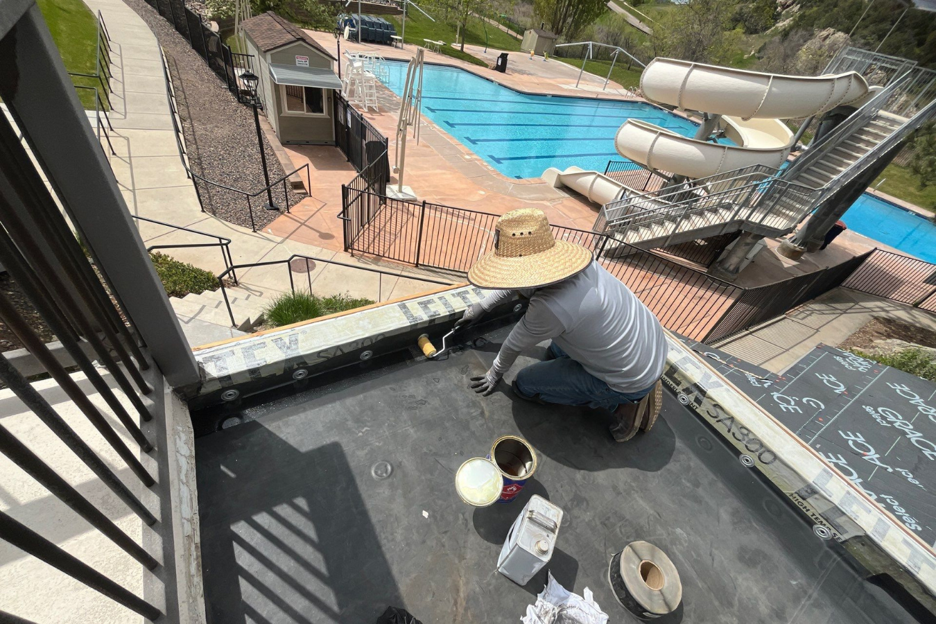 A worker wearing a large straw hat, gray long-sleeve shirt, and gloves is kneeling on a balcony, applying waterproofing to the edge of the roof near a swimming pool area.