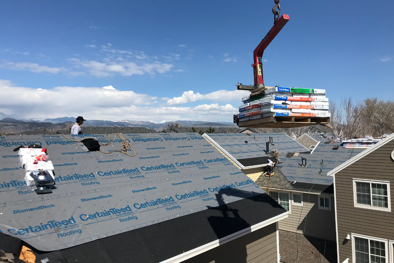 Roofers installing new roofing shingles on rooftops with a crane hoisting bundles of shingles in a suburban neighborhood.