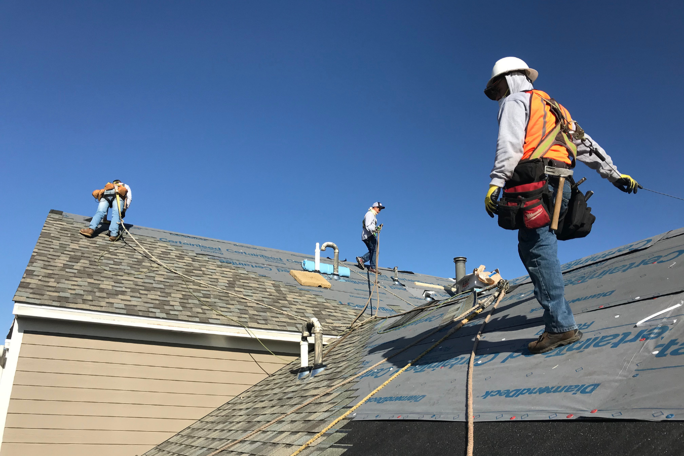 Construction workers repairing a roof on a house, using safety harnesses and ropes, under a clear blue sky.