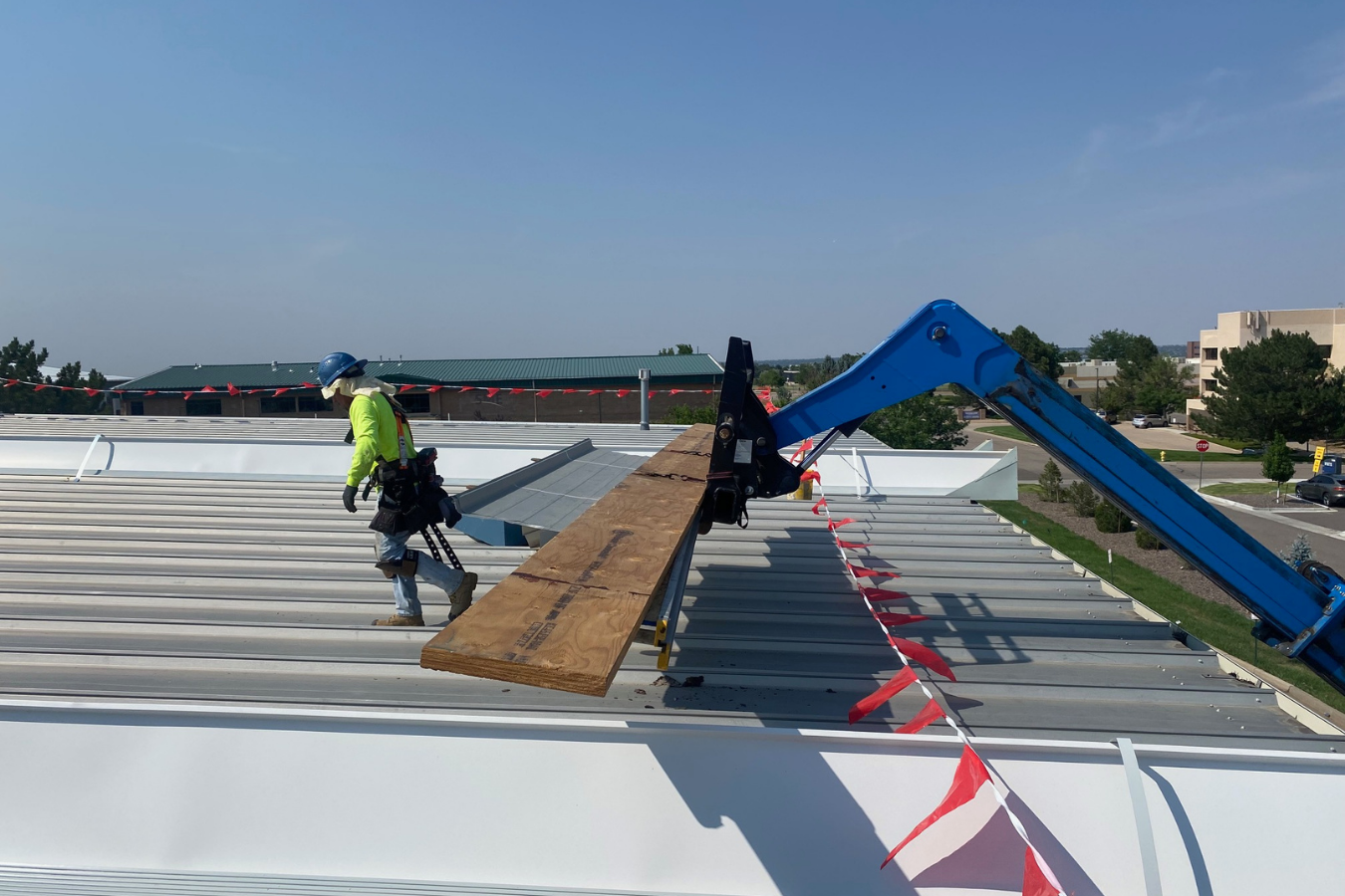 Construction worker on a metal roof with a blue crane lifting a wooden plank, safety barriers and trees in the background.