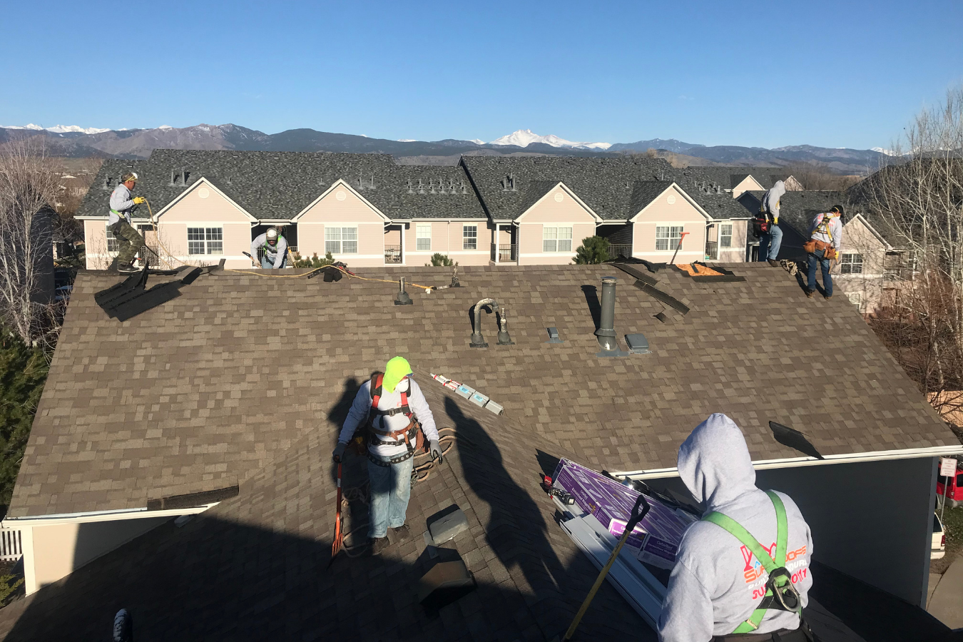 Roofers working on a house's roof, installing or repairing shingles under a clear blue sky with mountains in the background.