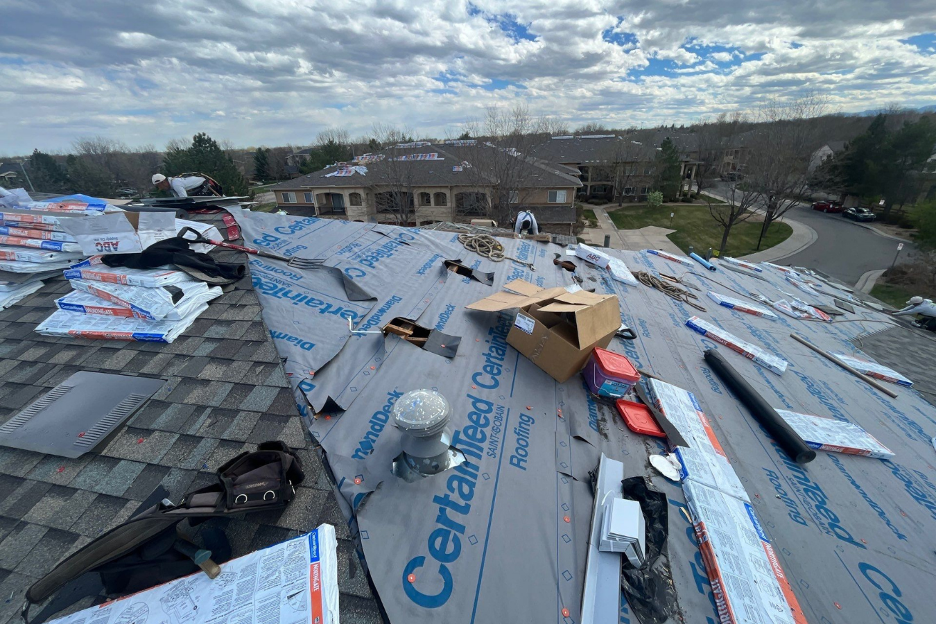 Residential roof in Colorado under construction with roofing materials, tools, and workers installing new shingles on a cloudy day.