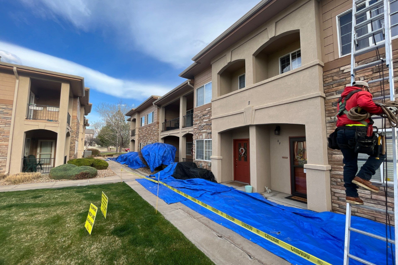 Construction worker climbing a ladder on apartment building, blue tarp on ground, caution tape, and warning signs
