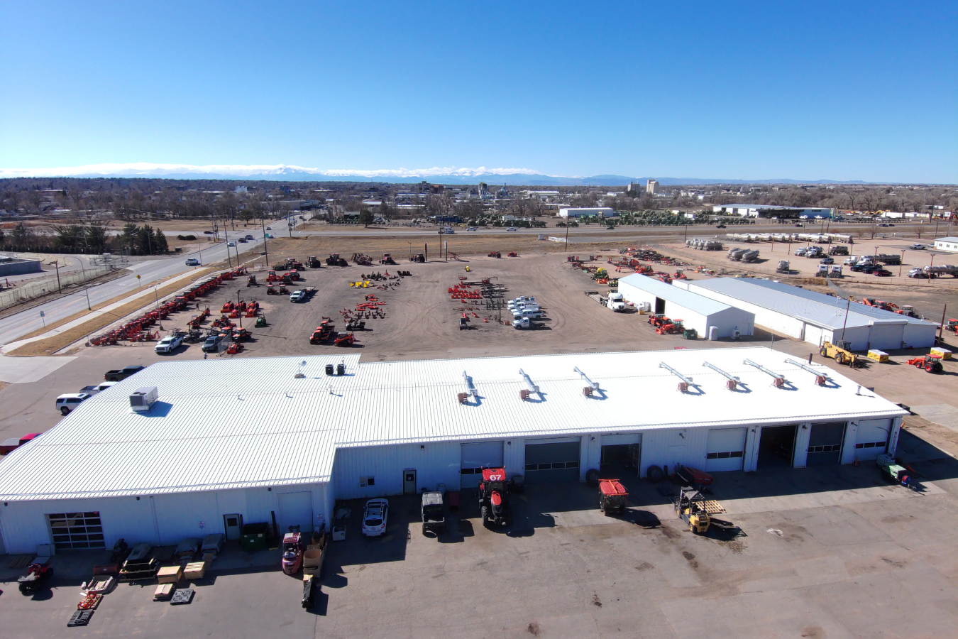 Aerial view of an industrial equipment store with a large white building and a gravel lot displaying various machinery and vehicles in a semi-rural area with mountains in the background.