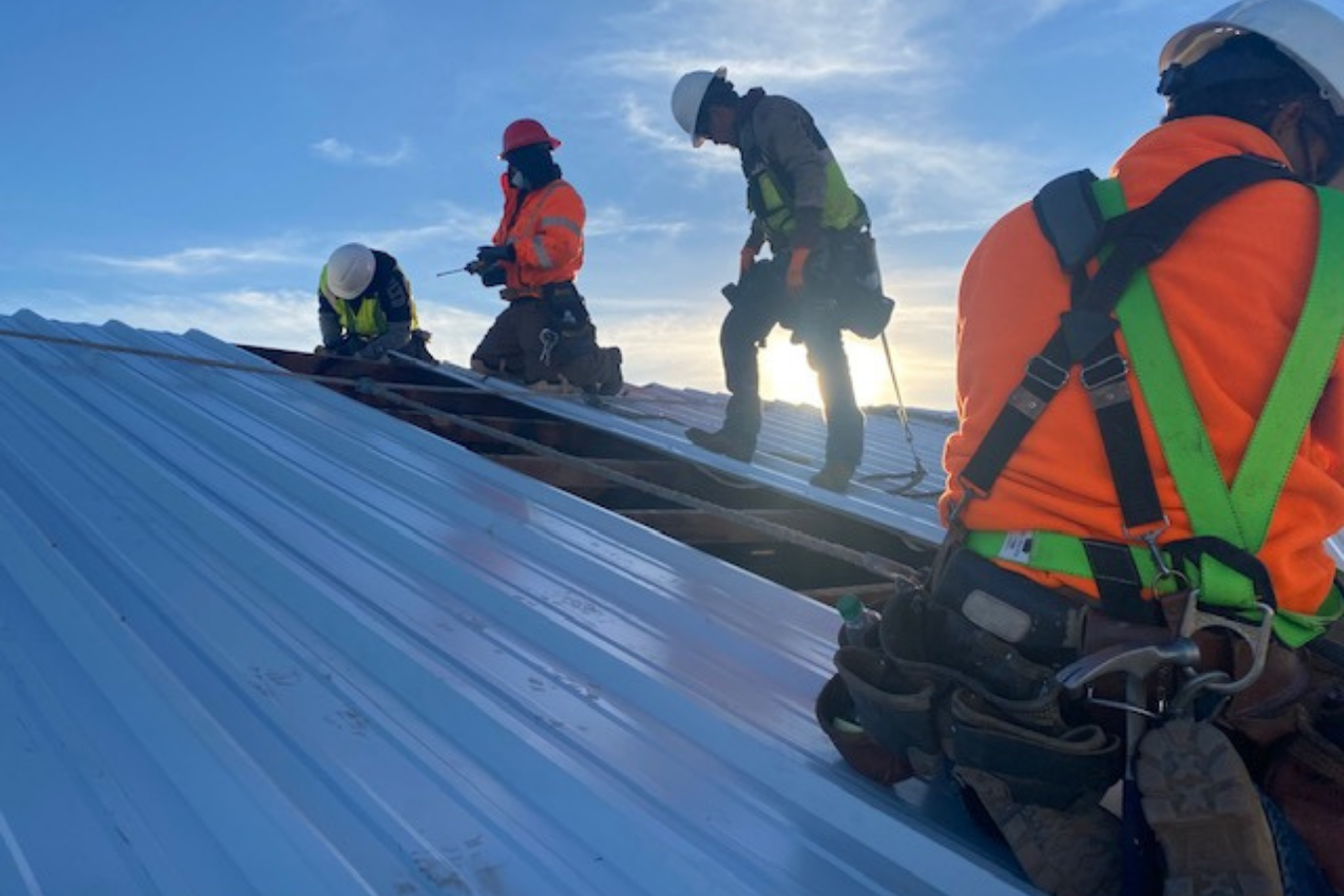 Construction workers on a roof with safety harnesses and helmets, during sunset.