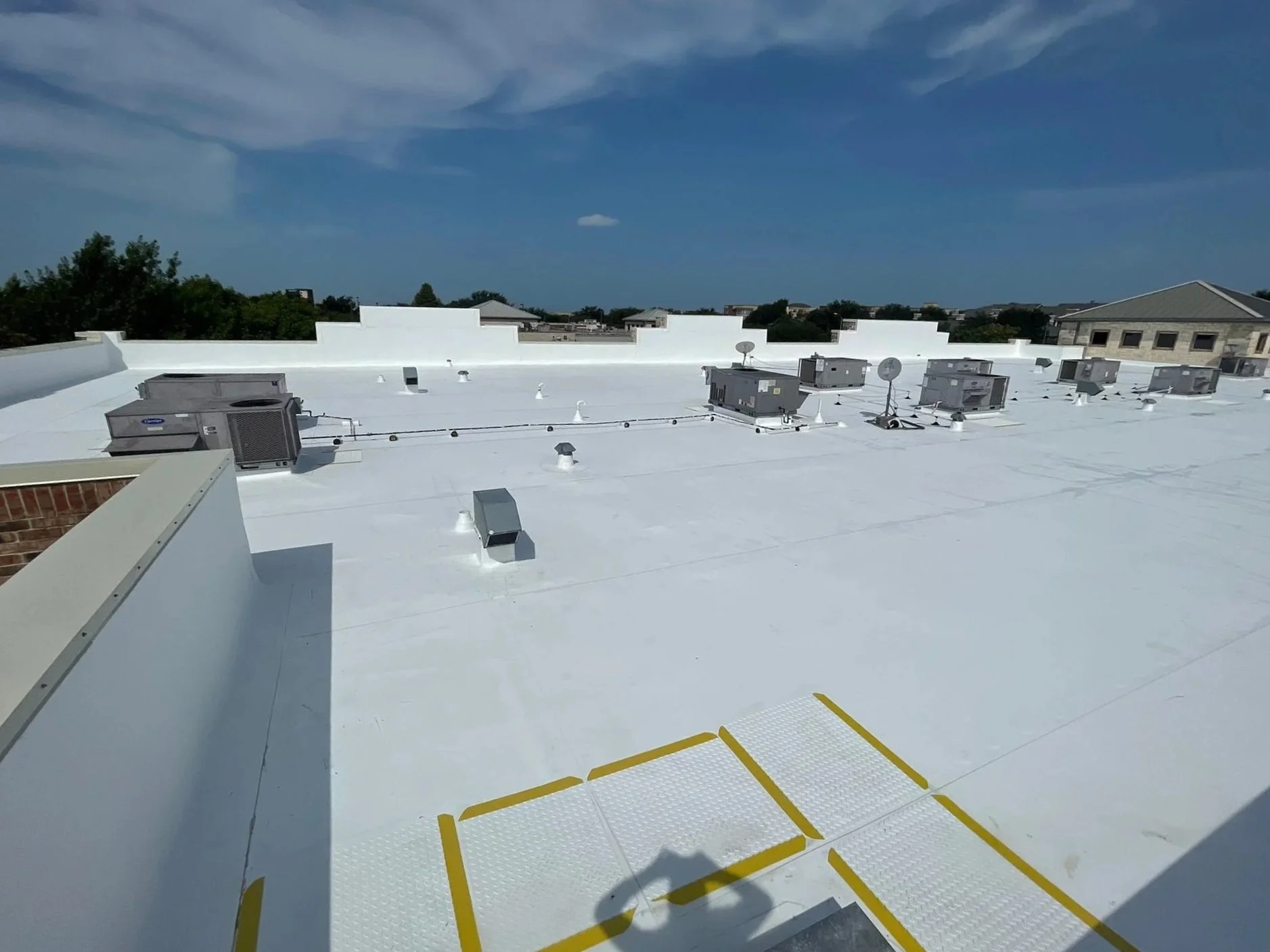View of a flat white commercial building roof with HVAC units, vents, satellite dishes, and yellow-marked equipment access areas, under a blue sky with some clouds.