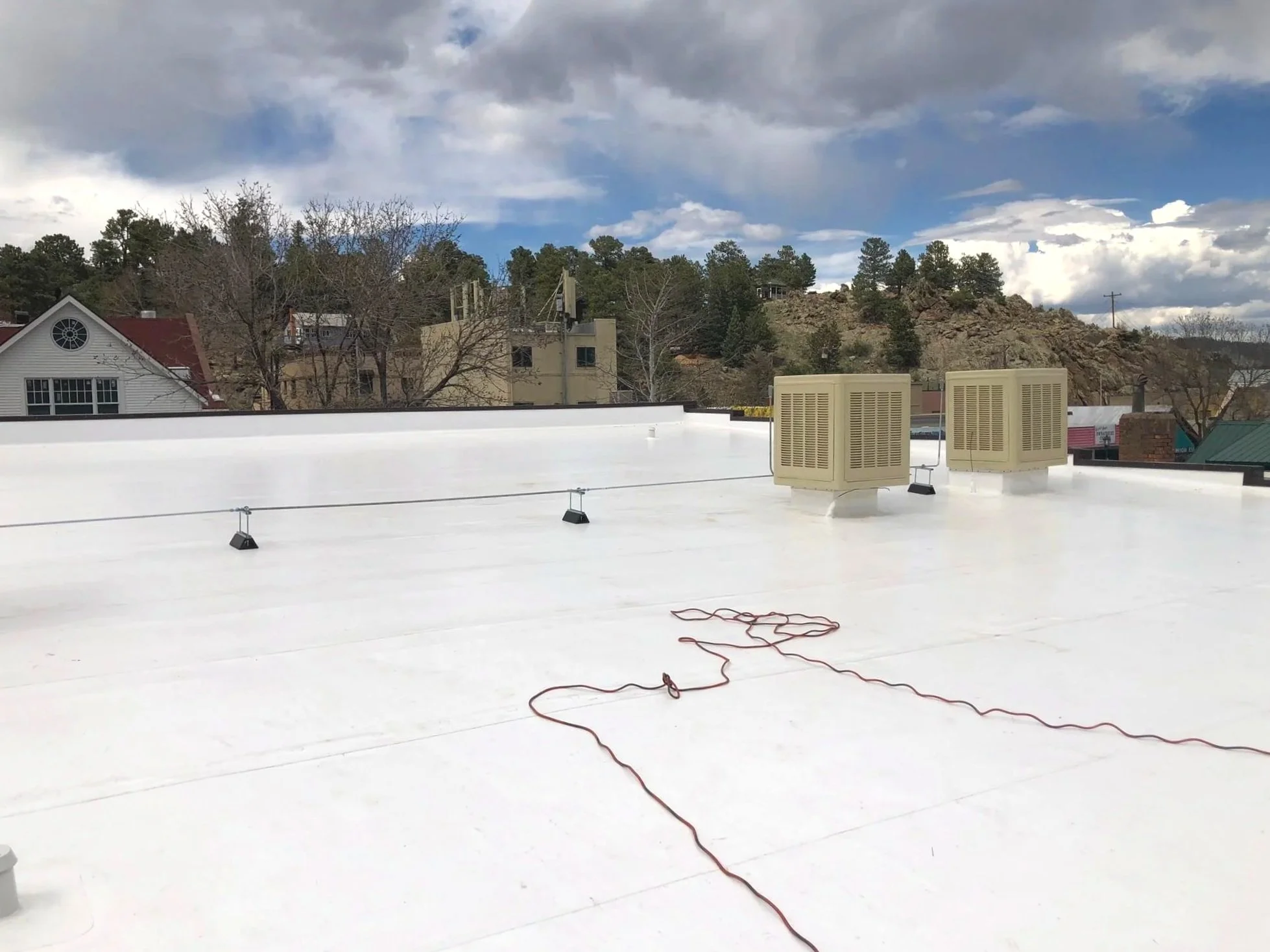 View of a flat white rooftop with two beige air conditioning units, an electrical cord, and small black lights attached to the roof. In the background, there are houses, trees, hills, and a partly cloudy sky.