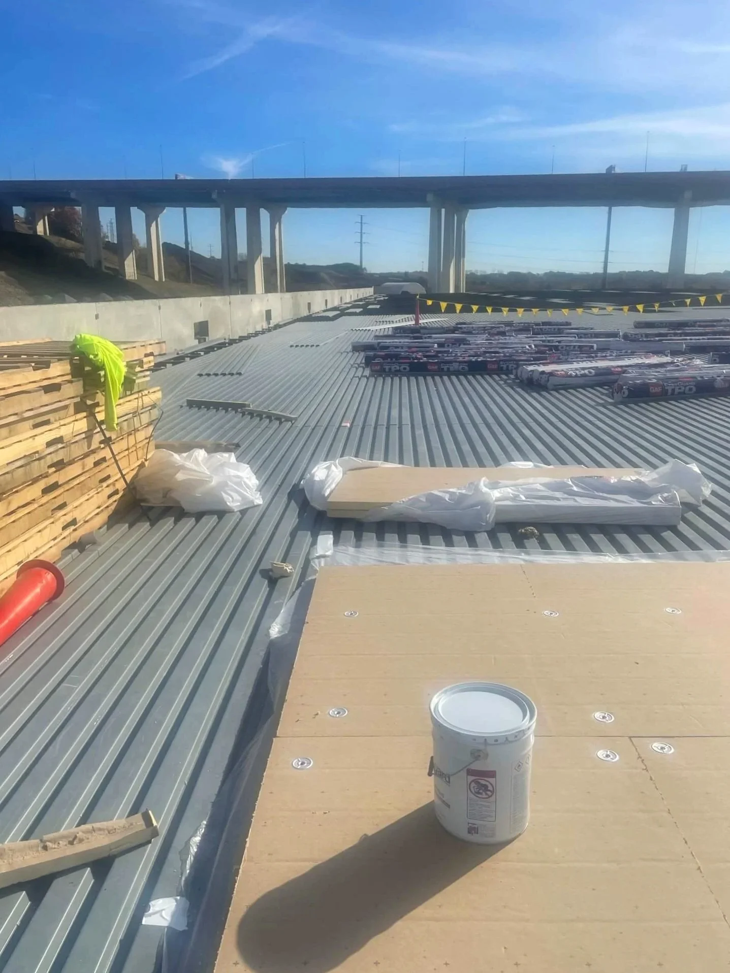 Construction site on a rooftop with steel roofing panels, wooden planks, a bucket of white paint, and safety equipment under a blue sky.