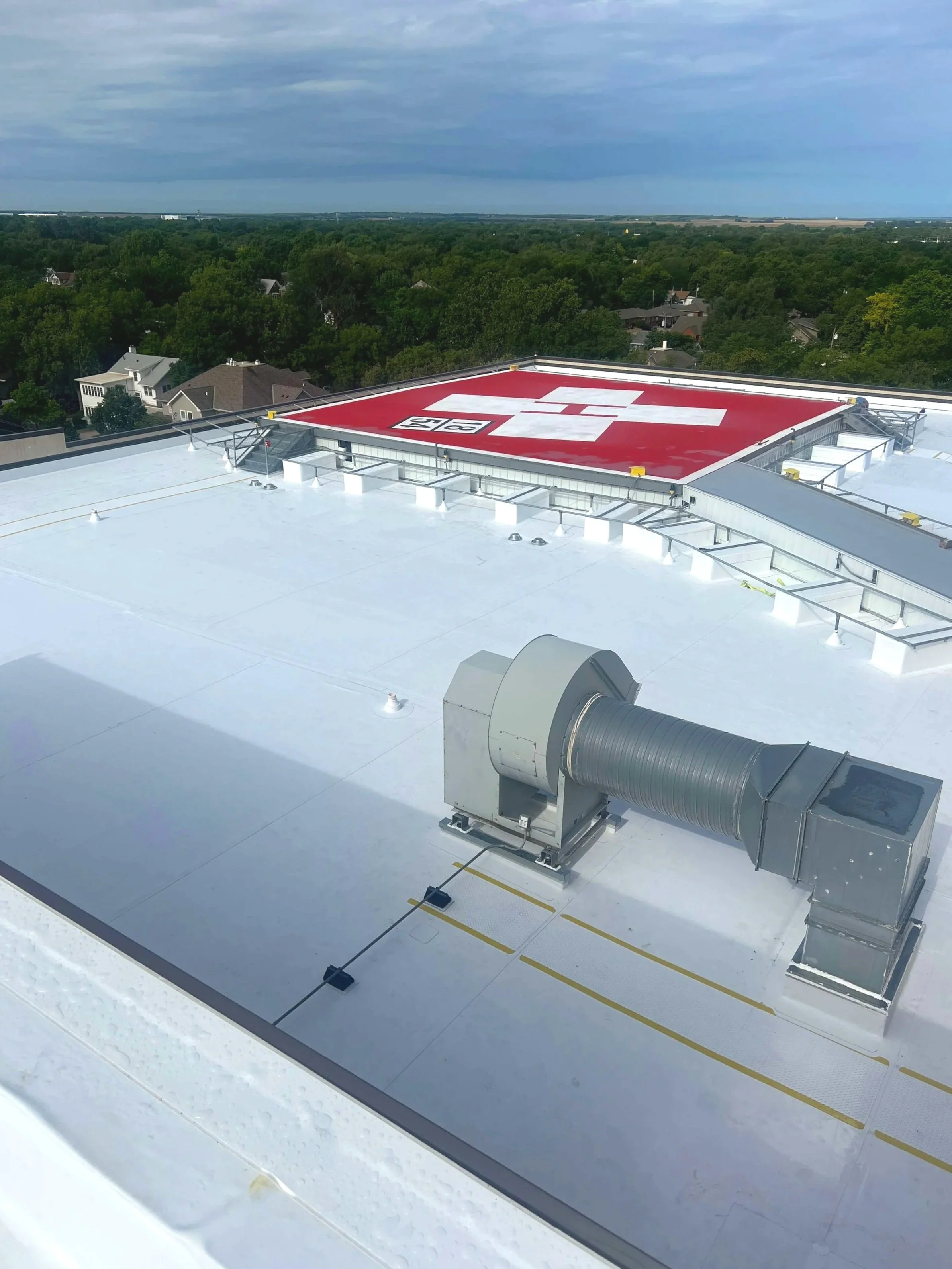 View of a rooftop with a red and white Swiss flag logo, a large HVAC ventilation system, and a cloudy sky with trees and houses in the background.