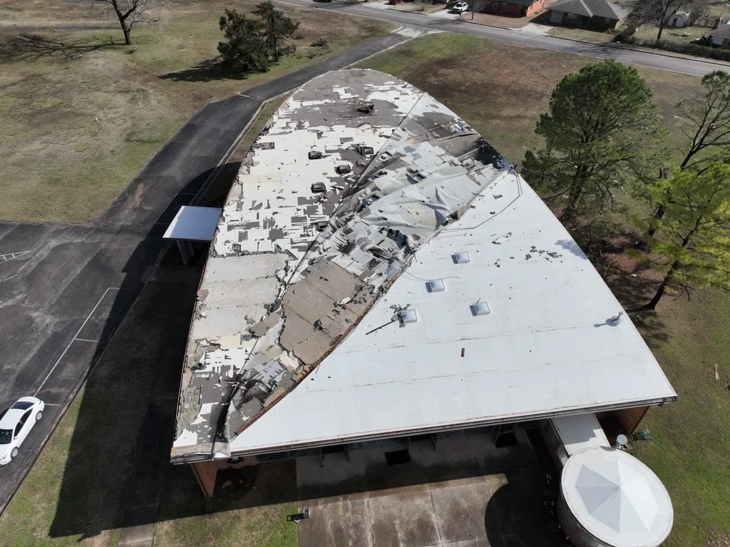 Aerial view of a building with a severely damaged, torn, and discolored roof after a storm or accident, surrounded by a parking lot, trees, and nearby residential houses.