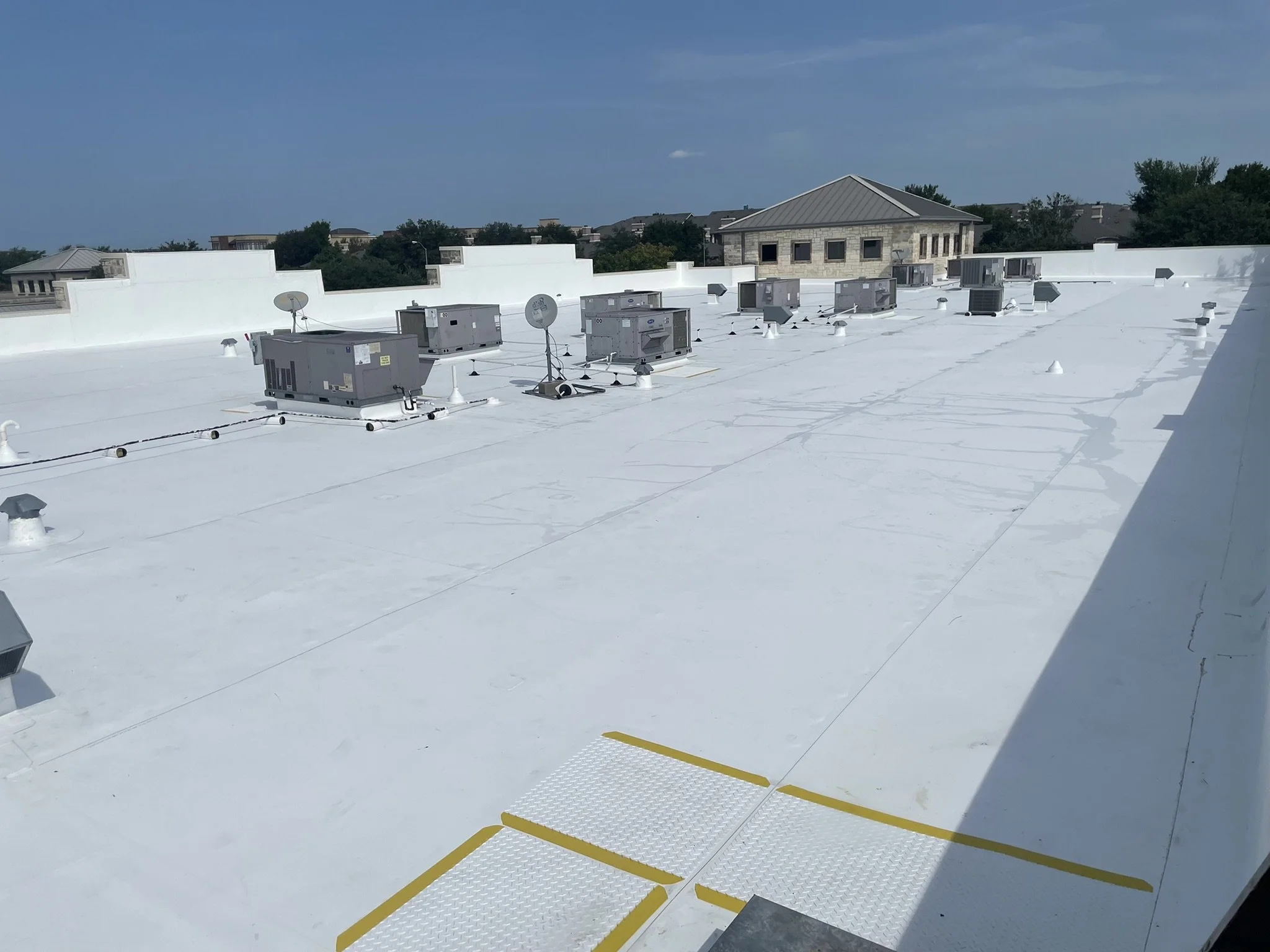 Flat white rooftop with HVAC units, satellite dishes, vents, and a yellow caution strip in the lower foreground, with a blue sky and trees in the background.