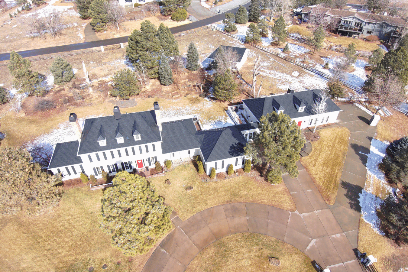 An aerial view of a large white house with a gray roof, surrounded by trees and a lawn, with some patches of snow on the ground.
