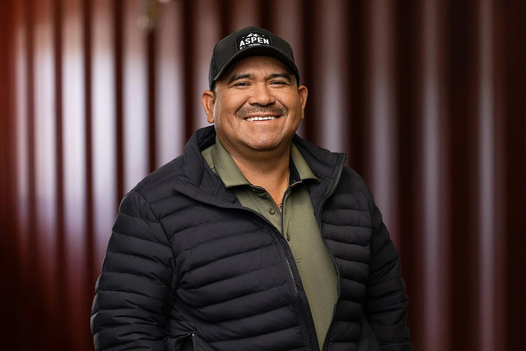 Everardo Moreno, wearing a black cap with the logo 'Aspen Colorado' and a black puffer jacket, standing in front of a blurred wooden background.