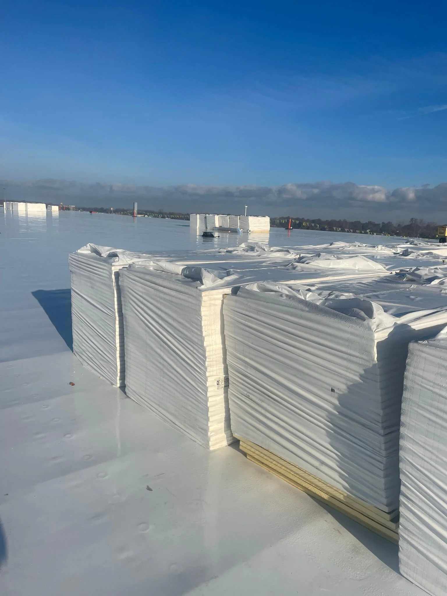 Large stacks of white insulation or construction material on a flat, snowy rooftop with a clear blue sky.