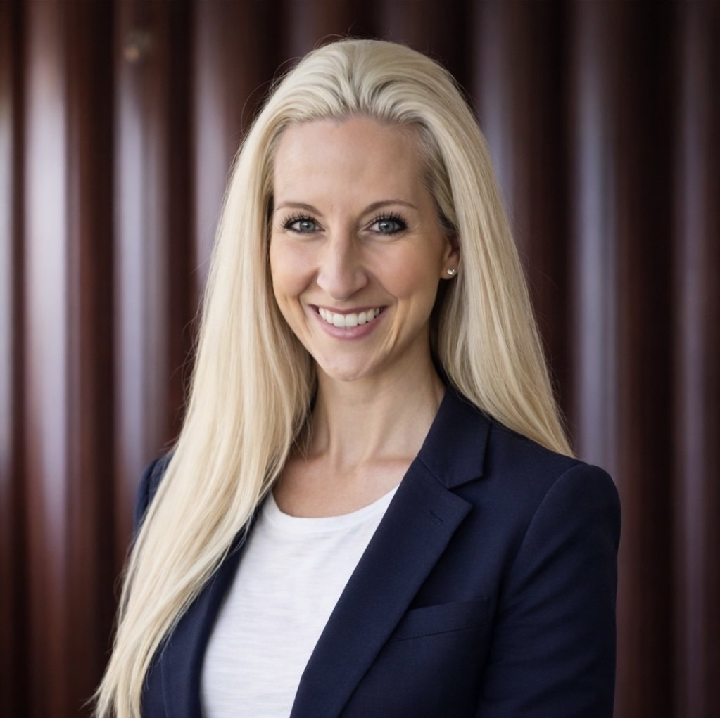 A woman with long blonde hair smiling, wearing a navy blazer and white top, against a dark wood-paneled background.