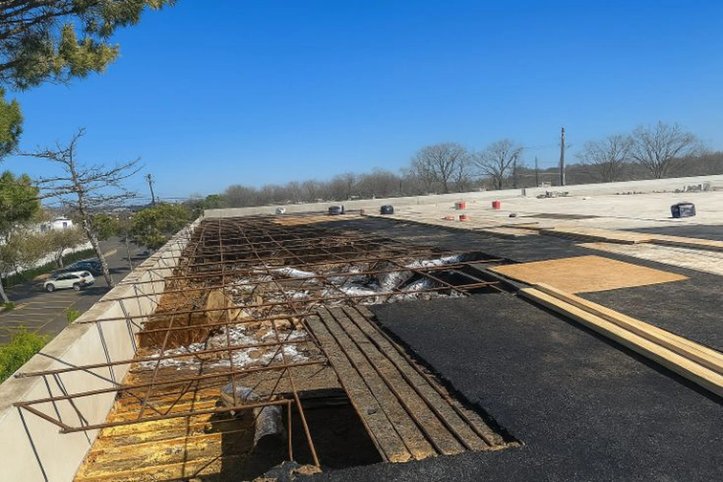 A rooftop under construction or repair, with exposed rusty metal rebar and plywood sheets, and some damaged sections, under a clear blue sky.