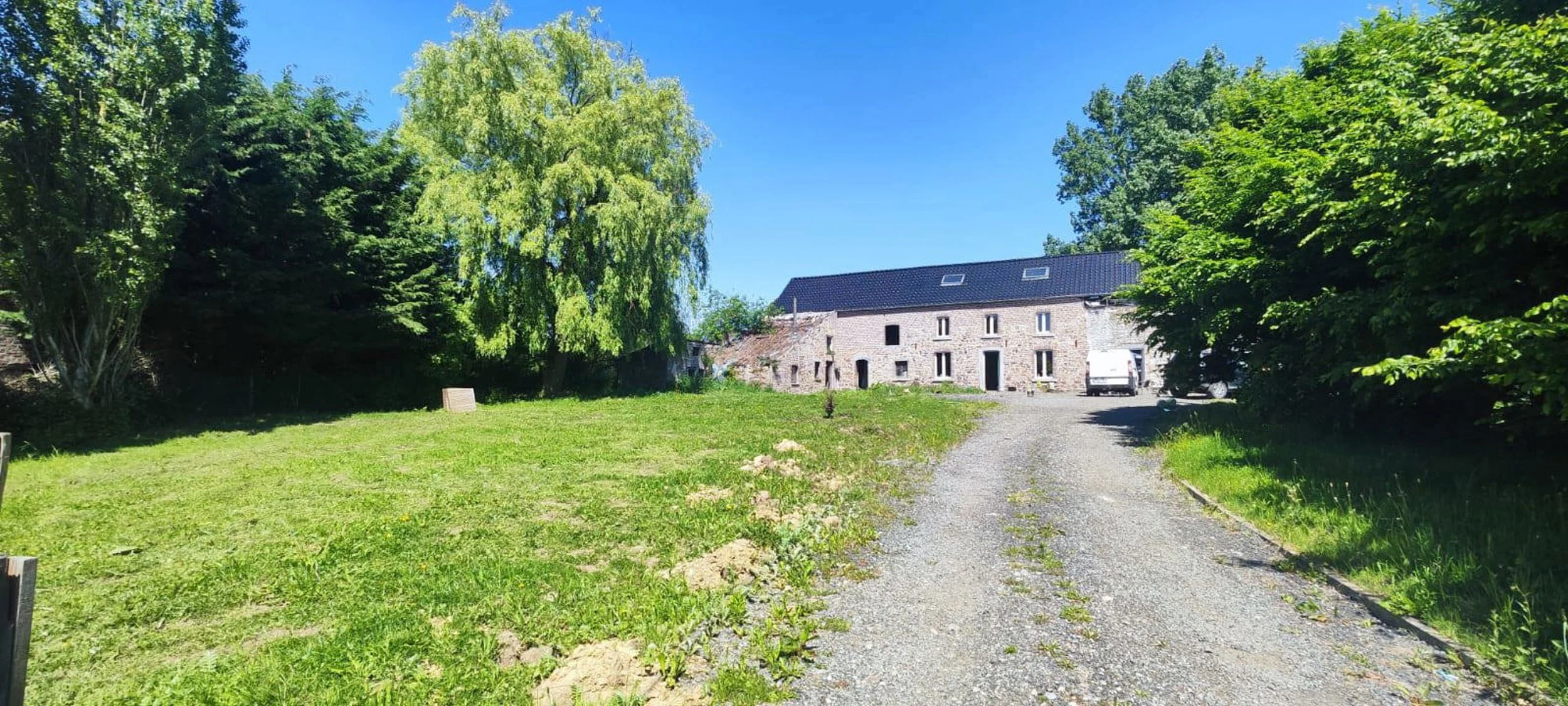 Une maison ancienne en pierre avec un toit en ardoise, entourée d'arbres verts, vue d'un chemin en gravier menant à la maison, sous un ciel bleu clair.