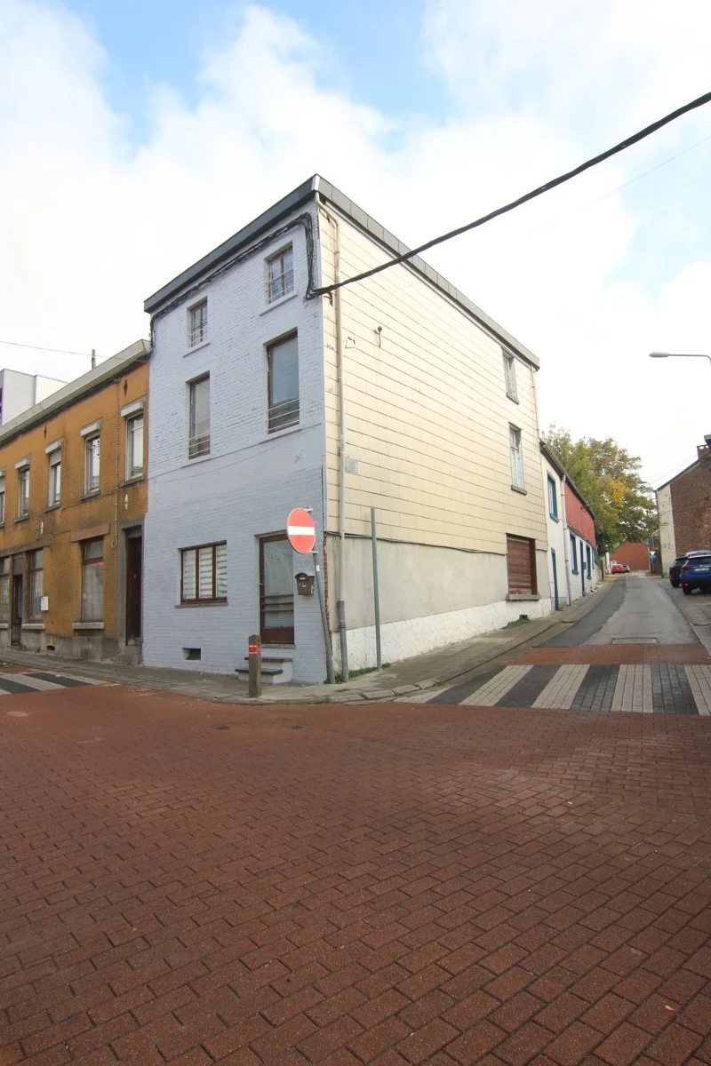 Une rue urbaine avec un bâtiment triangulaire à l'angle de la rue, des voitures garées et un passage piéton.