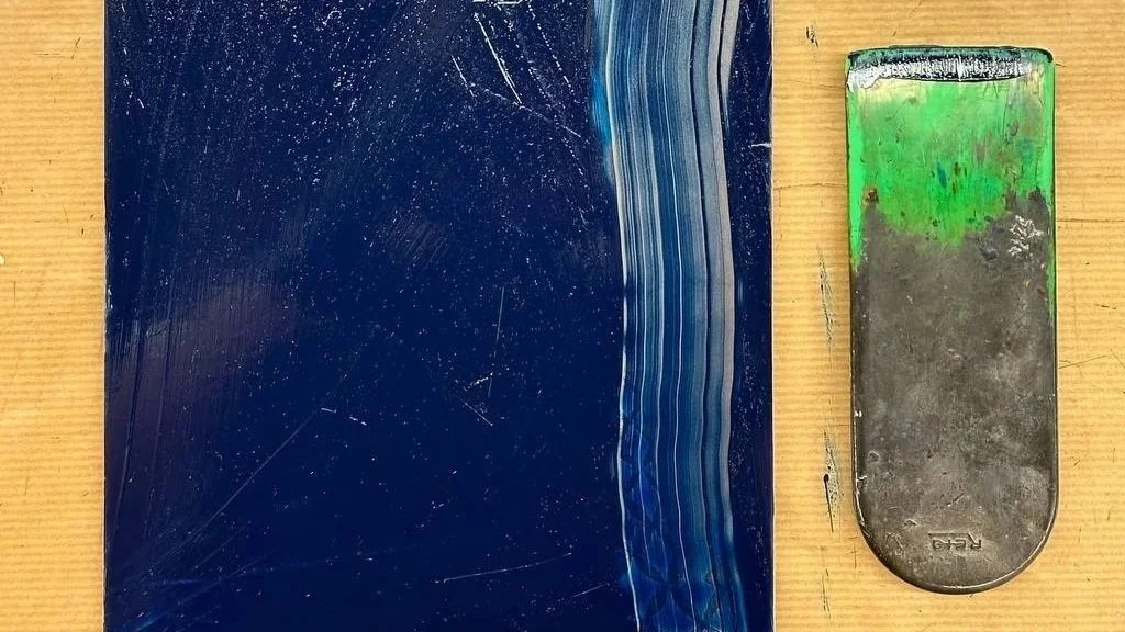 Una rasqueta de silicona ajada y una plancha de cobre cubierta con tinta azul y con rastros rayados, sobre una mesa de madera.