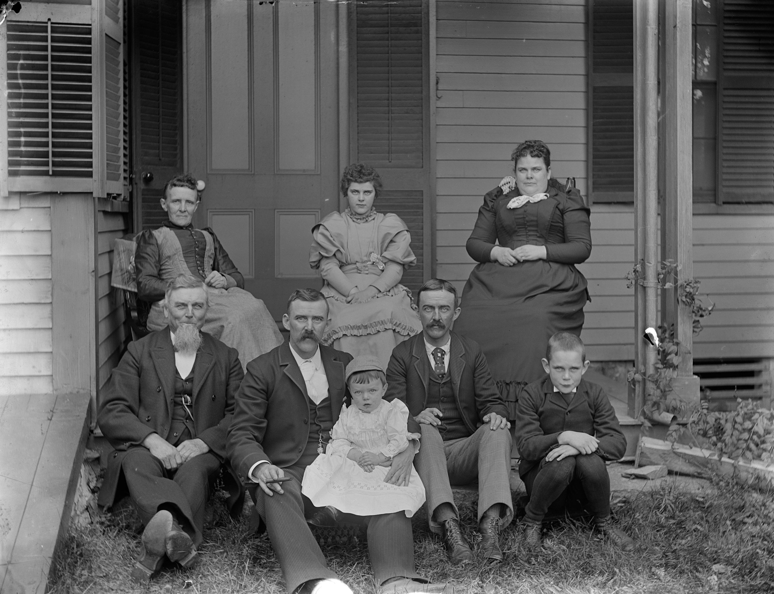 A black and white photo of nine people, including men, women, and children, posing on a front porch and steps of a house with wooden siding and a door with panels. The group appears to be family members from the early 20th century.