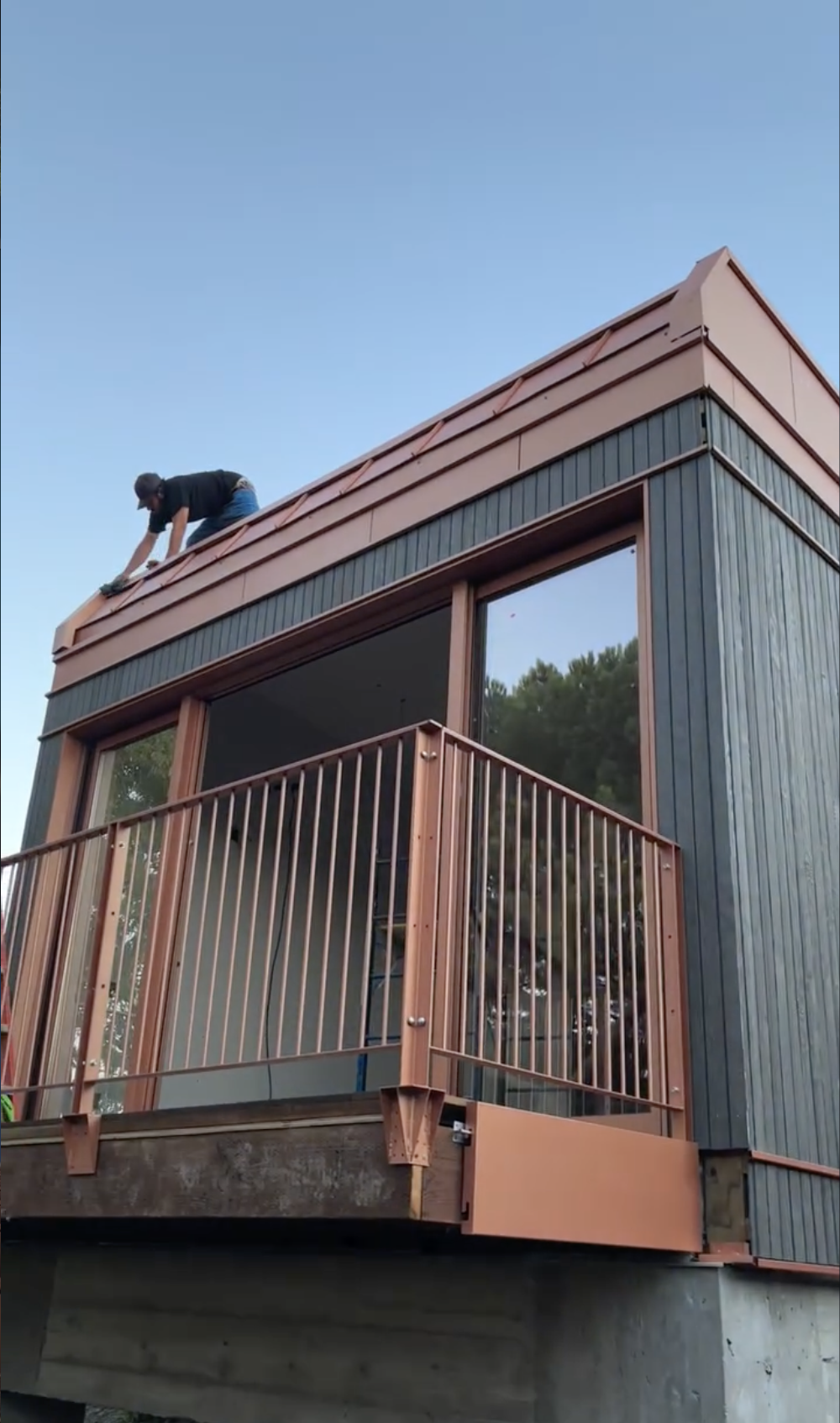 A person working on a sheet metal roof of a modern, multi-story building with black siding, large windows, and a balcony with a copper-colored railing in San Jose, CA