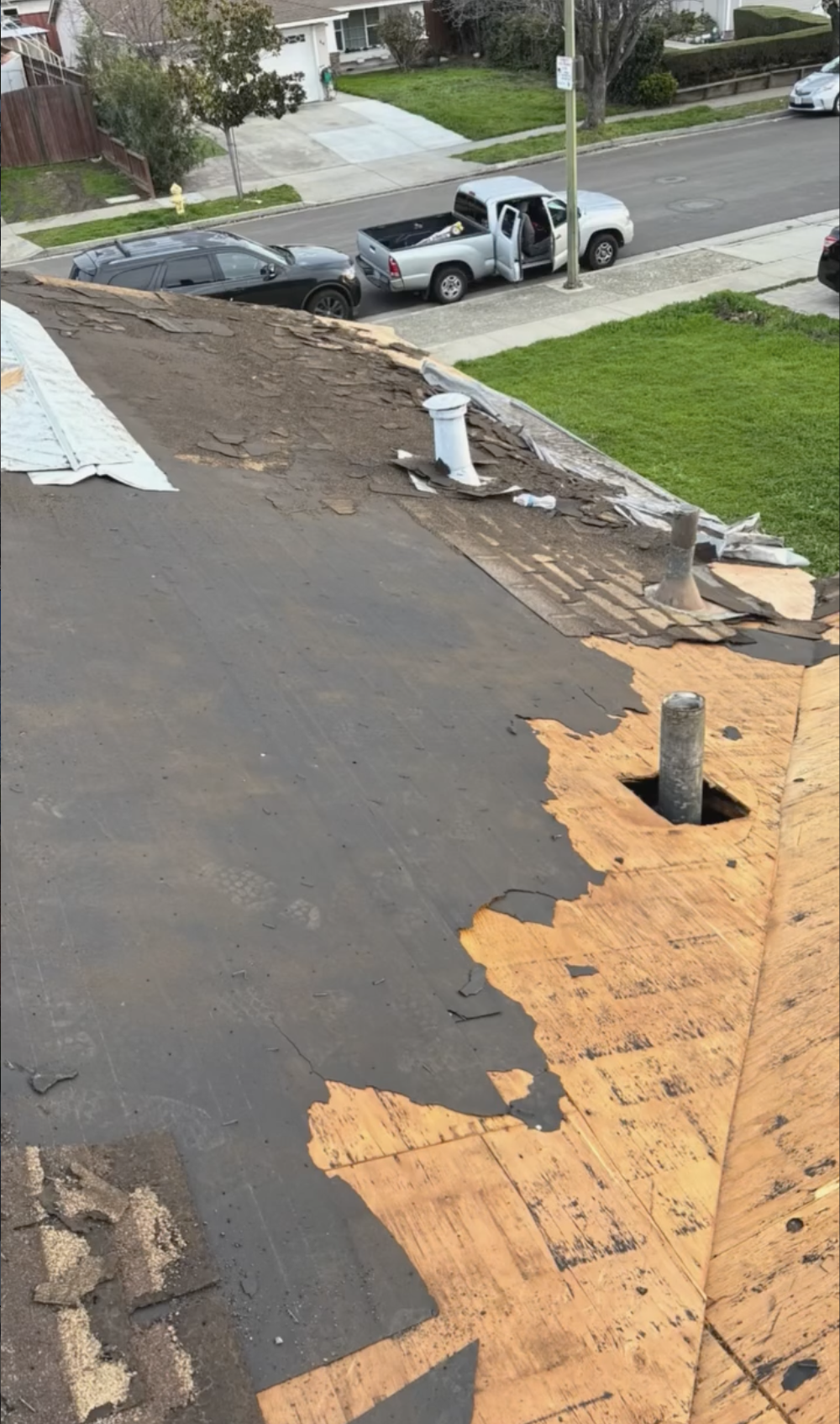 The roof is under construction, with damaged and peeling black shingles and exposed plywood. In the background, a street with parked cars and a white pickup truck with its driver’s side door open and items in the bed in Hollister, CA. 