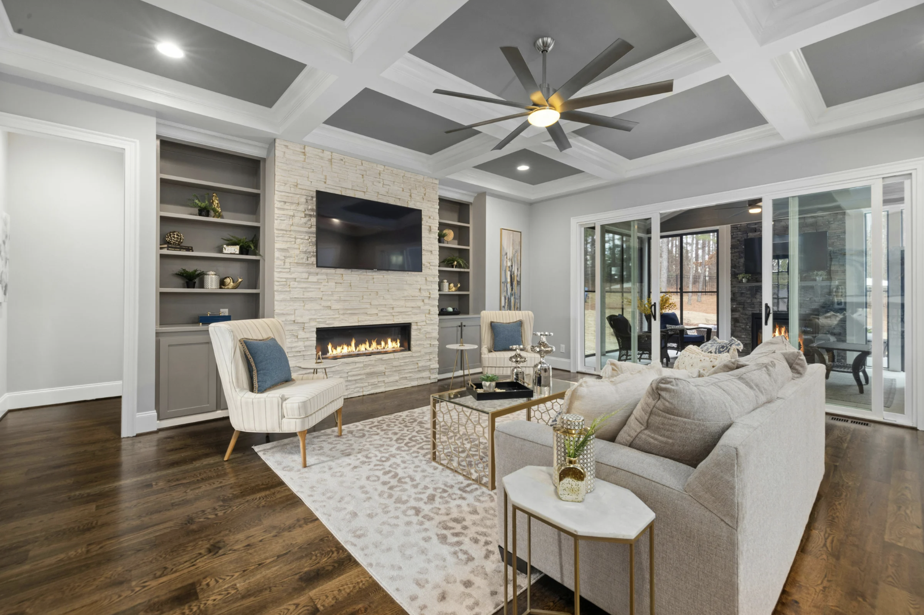 Modern living room with white painted walls, dark hardwood floors, a stone fireplace with a wall-mounted TV above, built-in bookshelves on either side, and a sliding glass door leading to an outdoor patio with a fire pit. The room features a beige sofa, a patterned area rug, a white armchair with blue pillows, and a gold-accented coffee table.