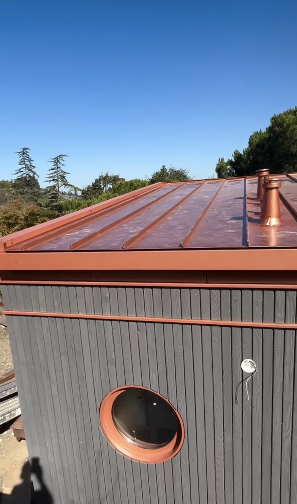 A building with a new installed copper-colored metal roof and gray wooden exterior, featuring a round window with a copper frame, under a clear blue sky and surrounded by trees in San Jose, CA.