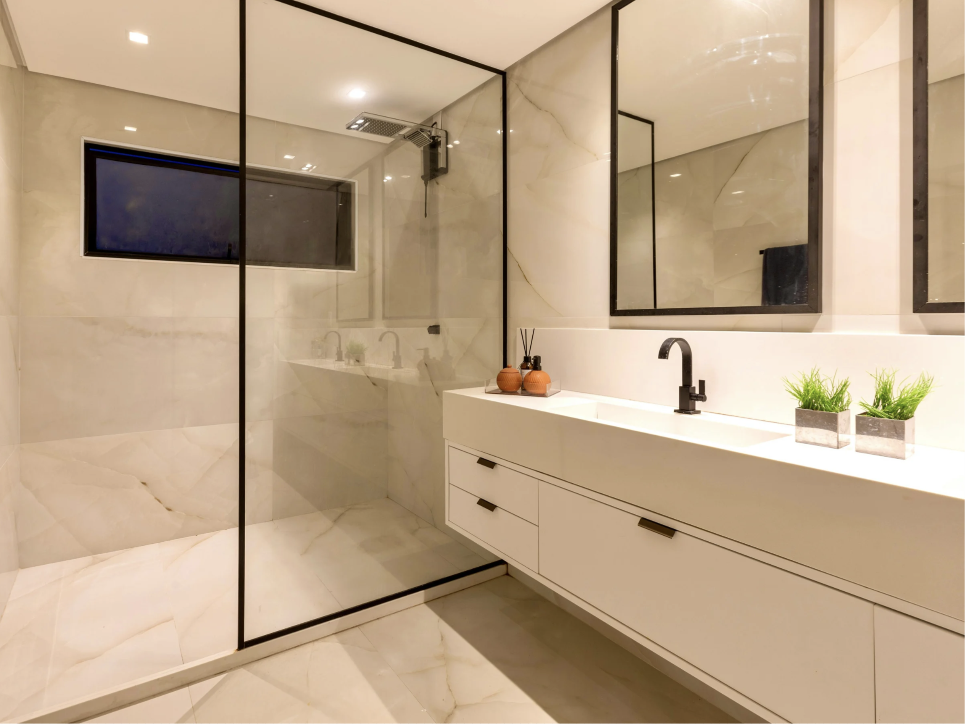 Modern bathroom with a glass shower enclosure, a white vanity with a black faucet, large mirrors, and decorative plants on the counter.