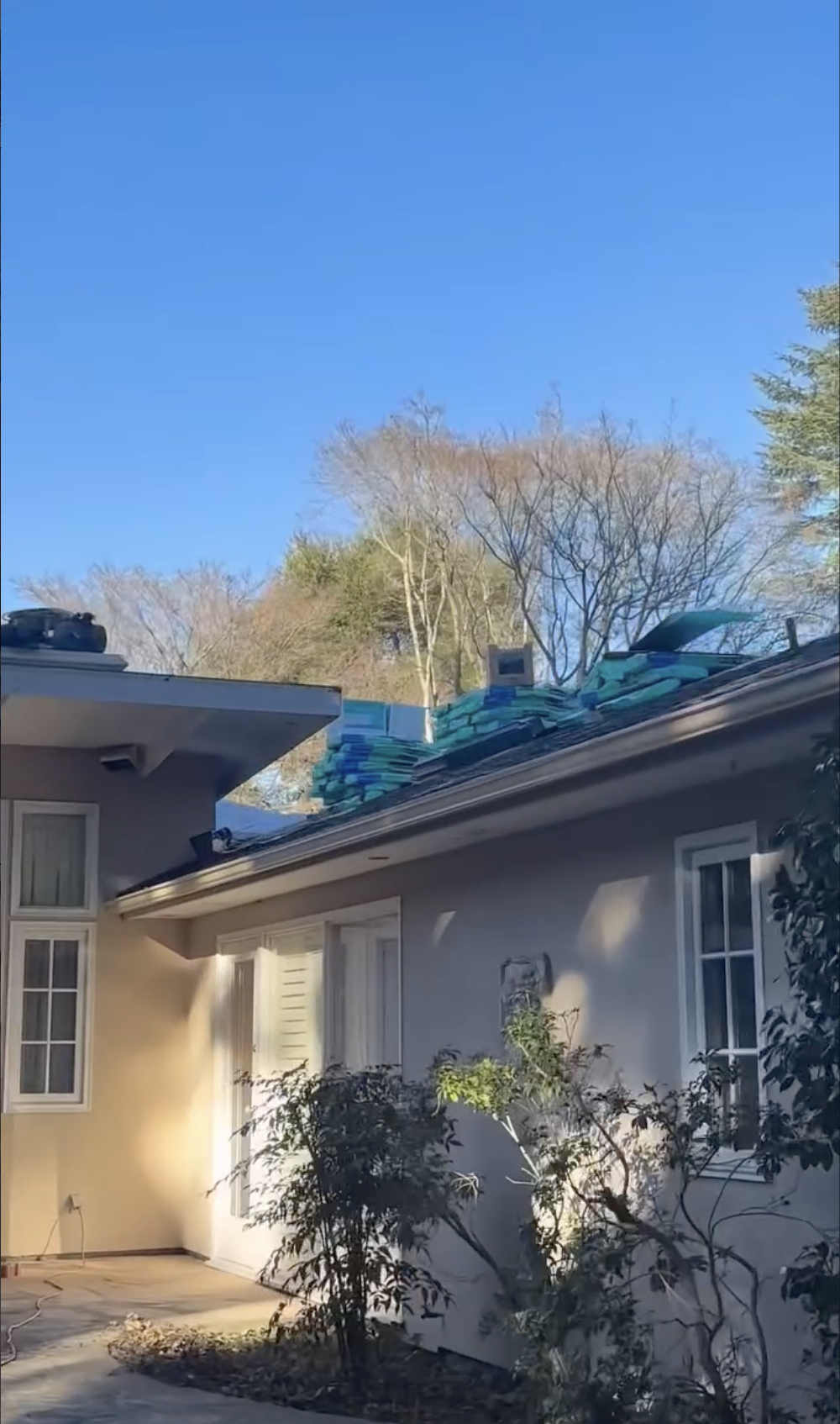 A house with a gray exterior, white windows, and plants in the front yard. The roof has a large stack of ice packs or insulation material on it, with some items placed on top in Gilroy, CA.