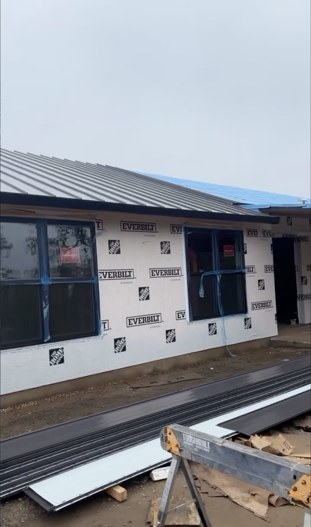 Construction site of a house with Everbilt building wrap, two open window frames partially covered with plastic, metal roofing sheets, and construction tools and materials on the ground in Hollister, CA. 