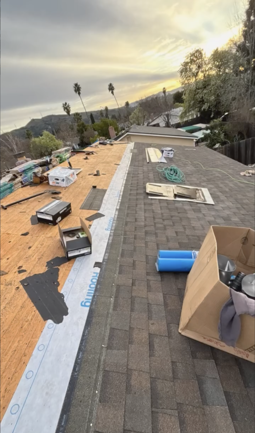 Rooftop under construction with tools, boxes, and materials scattered, showing partial new roofing installation at sunset, in a residential neighborhood with trees and hills in the background in San Juan Bautista, ca. 