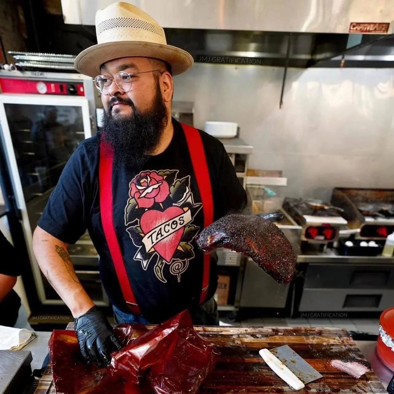 A man with a beard and glasses, wearing a straw hat and a black T-shirt with a tattoo on his arm, holds a smoked beef brisket in a restaurant kitchen.