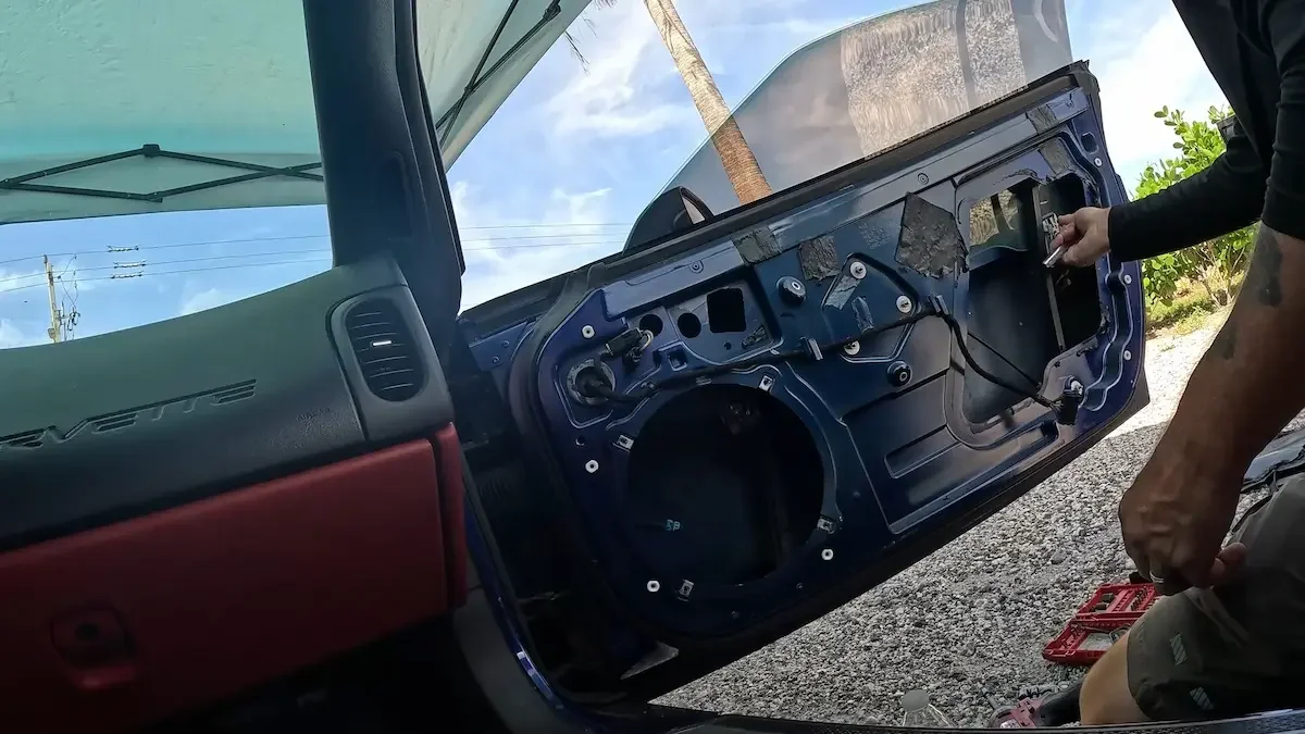 Person working on the inside of a car door, which is disassembled, showing mechanical components. The car is parked outdoors with a blue sky and some trees visible.