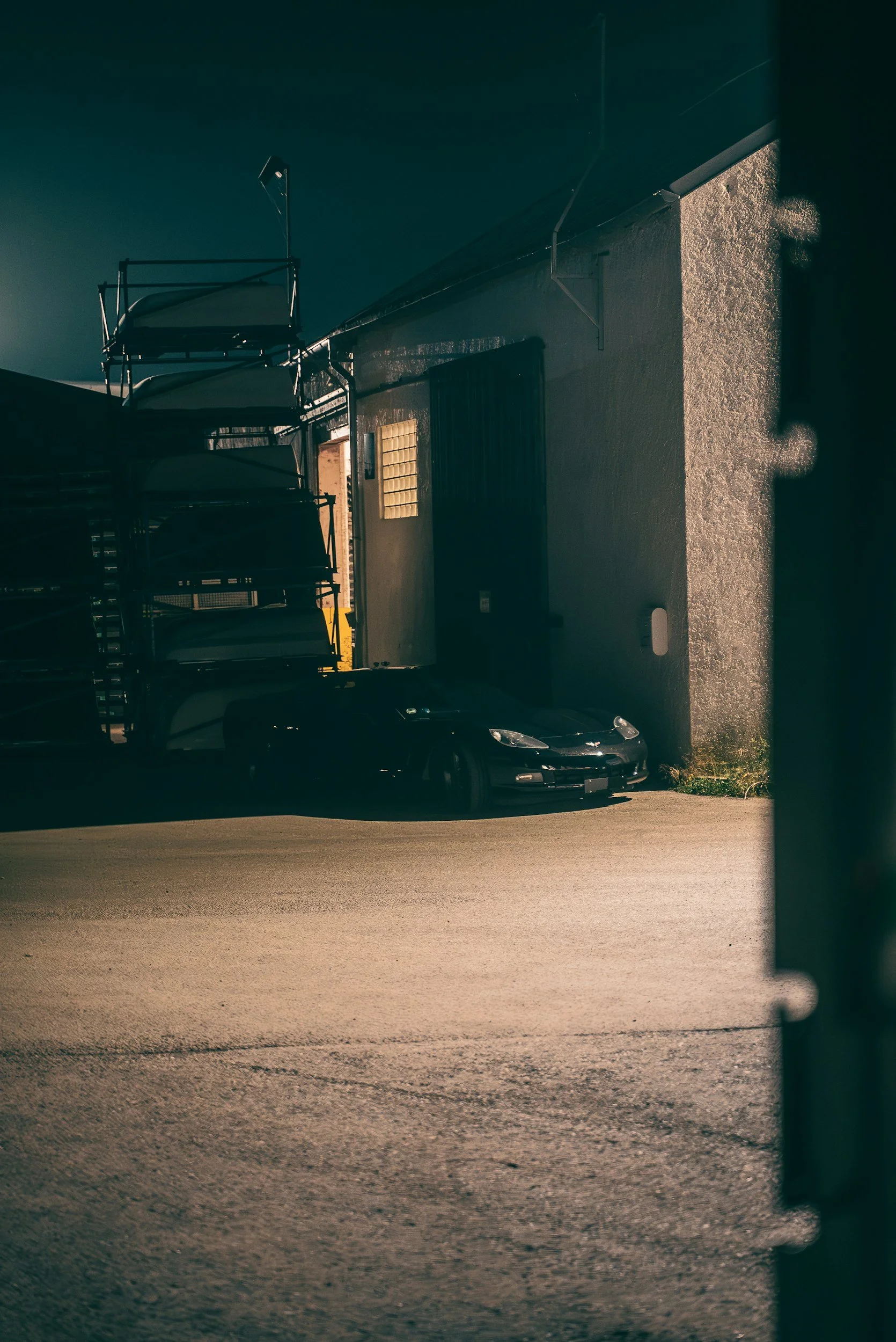 A dark-colored sports car parked near a building with a lit doorway, surrounded by stacked metal carts and a textured wall, illuminated by low light.