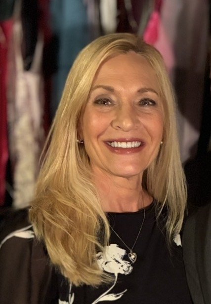 A smiling blonde woman with long hair, wearing a black top with a white floral pattern and a necklace, standing in front of a background with colorful clothing.