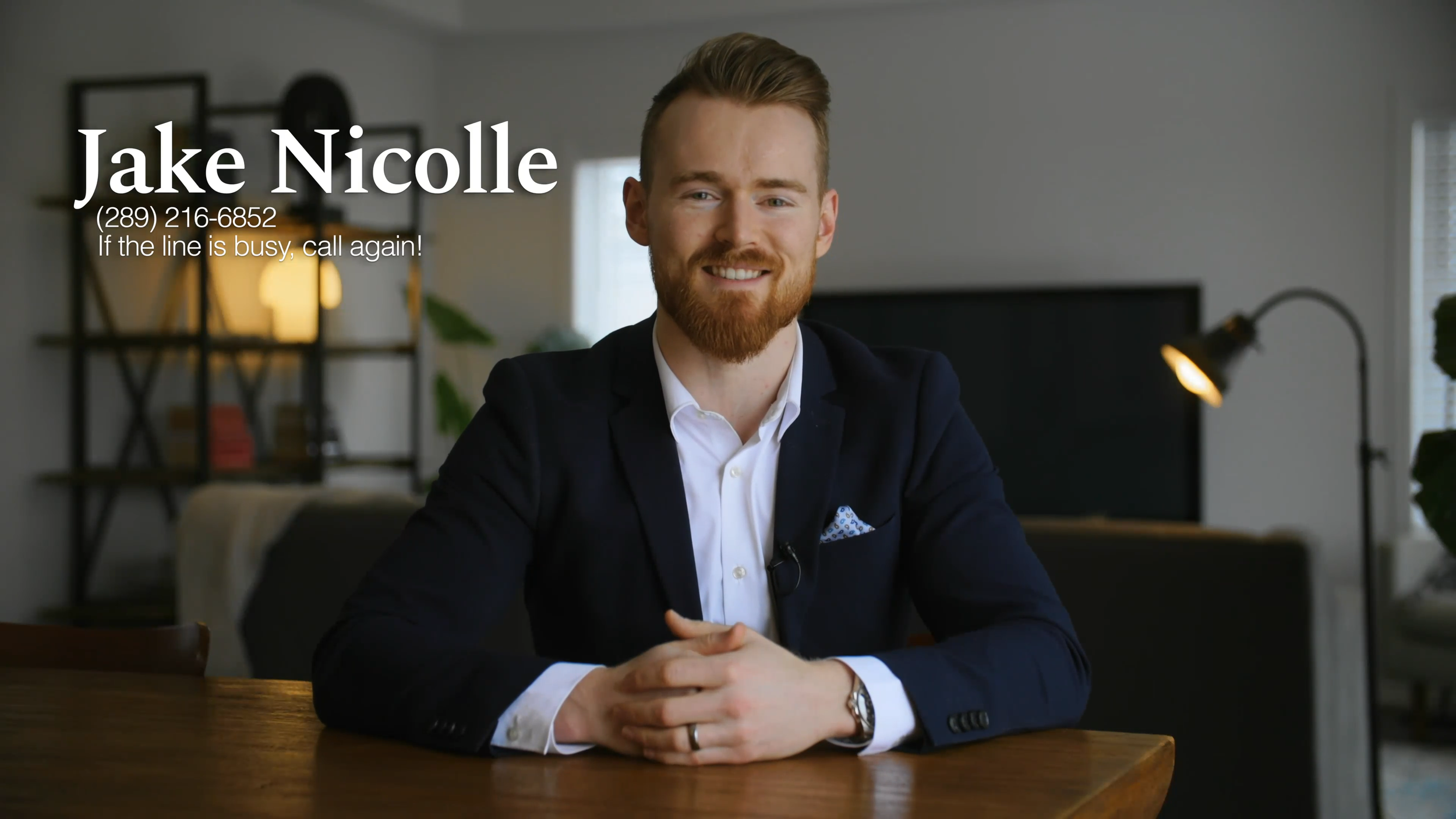 A man with red hair and a beard, wearing a dark suit and white shirt, sitting at a wooden table in a modern, well-lit living room with bookshelf, plants, and a lamp in the background.