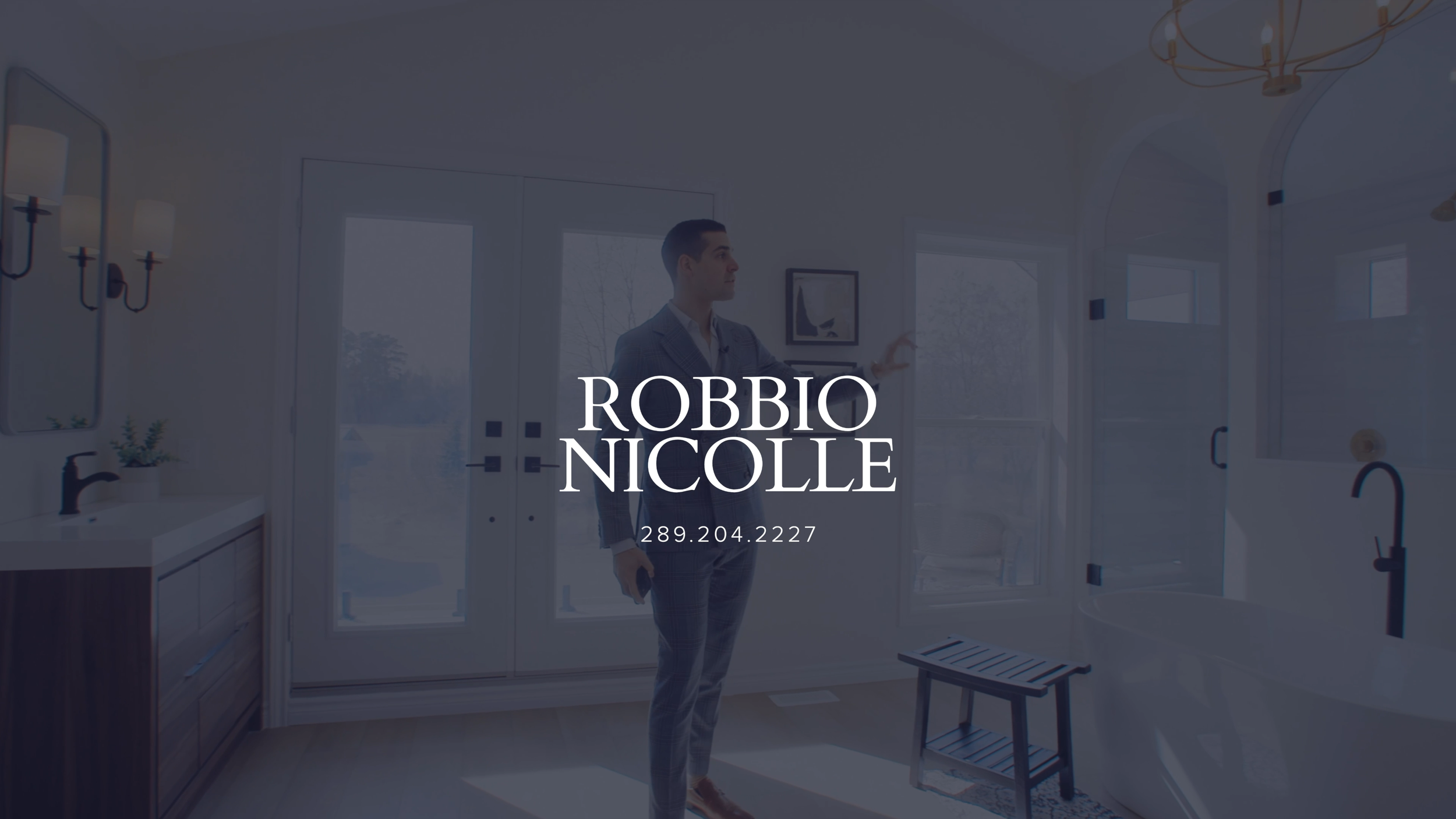 Man in a suit standing in a bright bathroom with large windows, a sink, mirror, bathtub, and artwork on the wall, holding a phone and gesturing with his other hand.